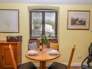 A dining area with a round wooden table set for two, a window with blinds, a framed map, and a framed painting on the wall. - Bradley Stables (Minehead)