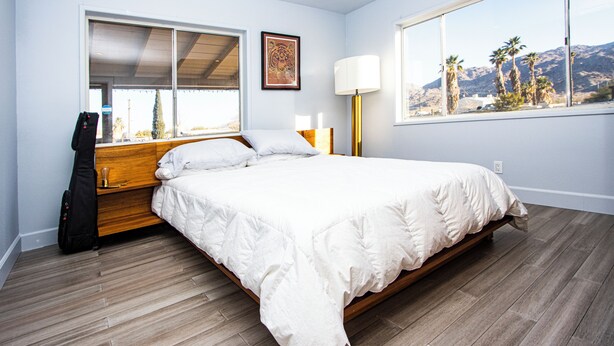 A bedroom with a large bed, a window with a view of palm trees and mountains, a guitar, and a lamp. - Experience the magic of Joshua Tree. Your cozy bungalow awaits. (Twentynine Palms)