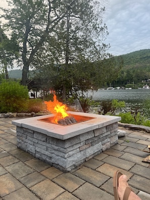 A stone fire pit with flames, surrounded by a stone patio, near a lake with boats and trees.