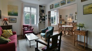 Un salon chaleureux avec un canapé, des fauteuils, une table basse et une bibliothèque. De grandes fenêtres laissent entrer la lumière naturelle et offrent une vue sur la verdure environnante.