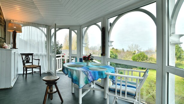 Un espace extérieur abrité, aménagé pour les repas, avec une table pour deux personnes, des chaises blanches et une chaise dont l’assise est en osier tressé.