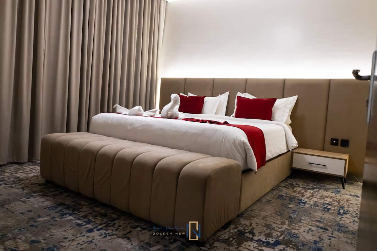 A hotel room with a large bed, a bedside table, a footstool, and a carpeted floor.