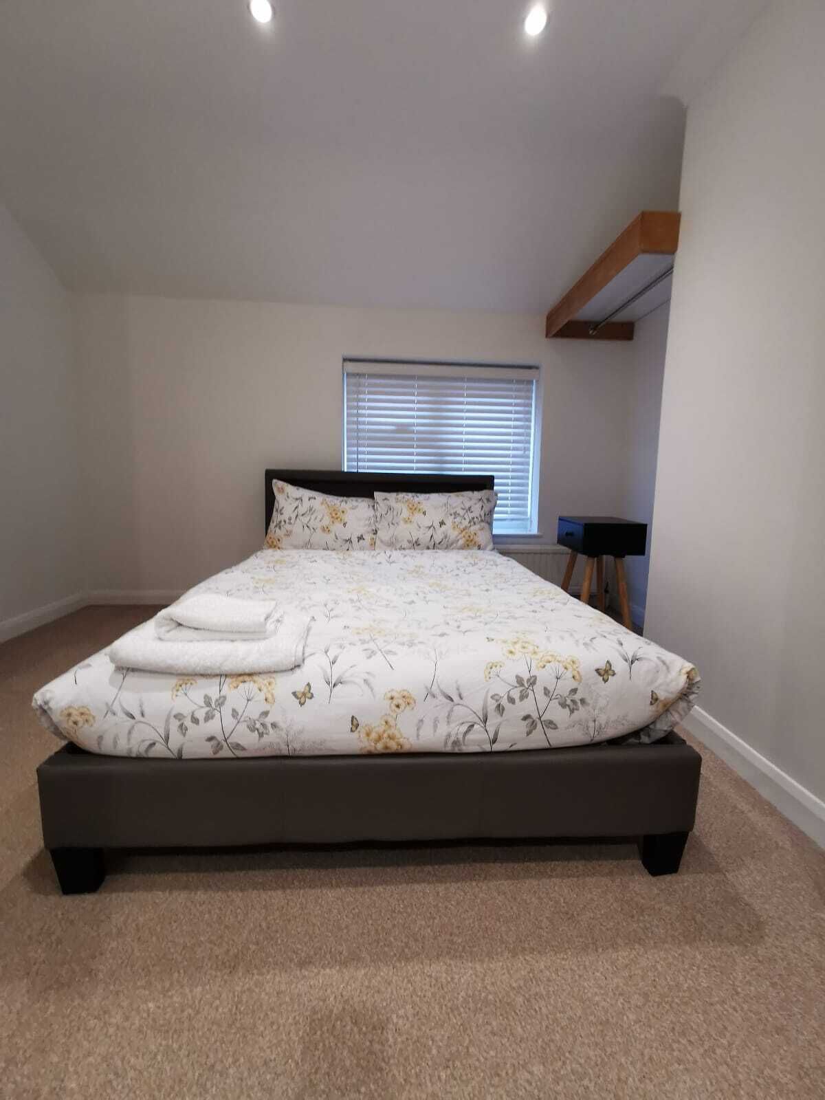 A bedroom with a large bed, a window with blinds, a small table, and a wooden beam on the ceiling.