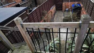 A wooden deck with a metal railing, overlooking a fenced backyard with a brick wall and a blue container.