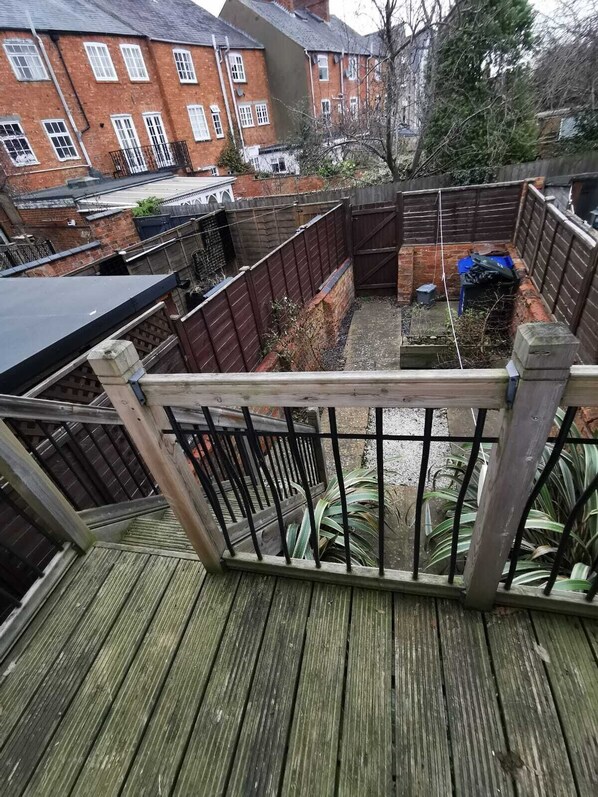 Eine Holzterrasse mit Metallgeländer, die auf einen umzäunten Garten mit Backsteinmauer und einem blauen Container blickt.