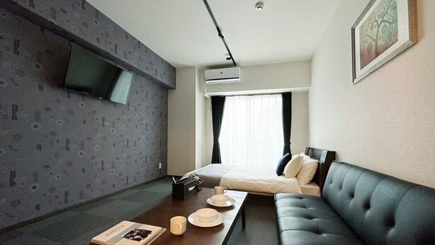 Une chambre d’hôtel moderne dotée d’un téléviseur à écran plat fixé au mur, d’un lit avec une tête de lit sombre, d’un canapé en cuir noir et d’une table basse en bois sur laquelle se trouvent un livre et une tasse.