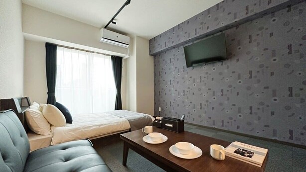 A modern hotel room with a sofa, a bed, a dining table with plates and a cup, a flat-screen TV mounted on the wall, and an air conditioning unit. - Nagomi by b hotel | Comfortable Studio Stay N62 (Hiroshima City)