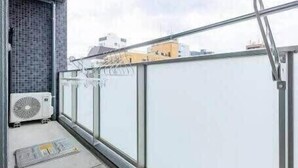 Un balcón con barandilla de cristal, un equipo pequeño de aire acondicionado y vista a edificios en la distancia.
