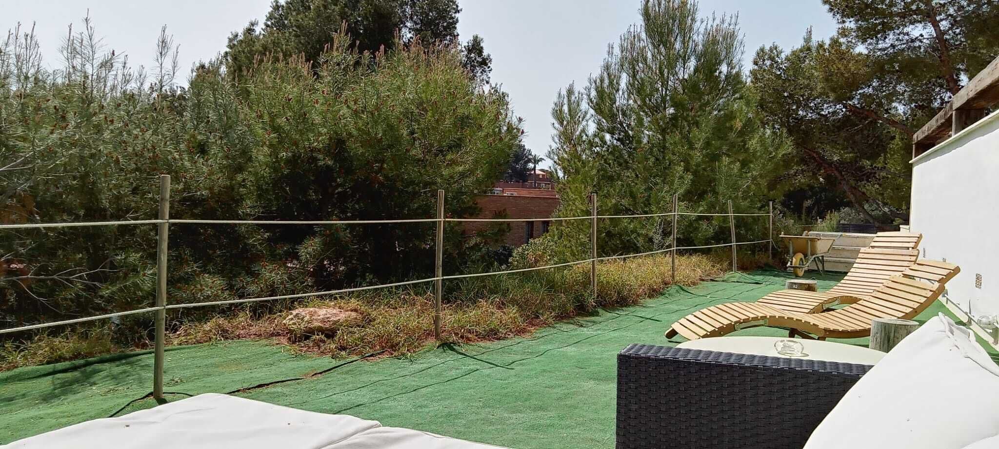 A rooftop terrace with wooden lounge chairs, a green artificial turf, and a view of surrounding trees and a building.