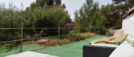 A rooftop terrace with wooden lounge chairs, a green artificial turf, and a view of surrounding trees and a building.