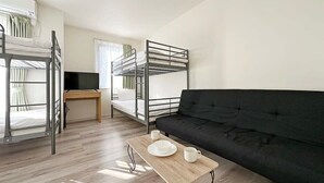 Una habitación tipo dormitorio con literas, un sofá negro, una mesa de madera con una taza y un platillo, y un televisor sobre un escritorio.