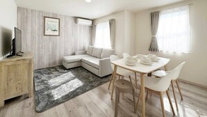 A modern living room with a sofa, a dining table with chairs, a TV on a wooden cabinet, and an air conditioning unit on the wall. - Spacious 2BR Apartment very near the Station (Onomichi)