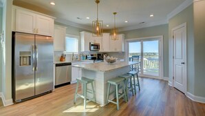 A modern kitchen with white cabinetry, stainless steel appliances, and a central island with stools. - Flamingo Villa - Oceanfront B Luxury Home, Hot Tub, 6 bed 4.5 bath (North Topsail Beach)