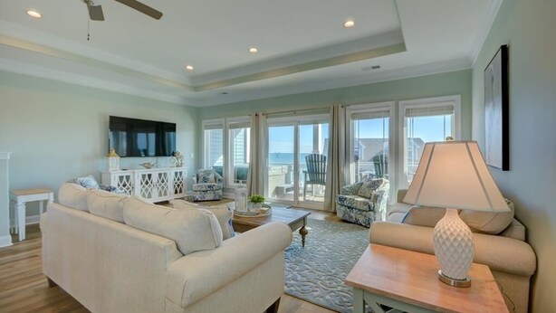 A spacious living room with a large sofa, a coffee table, and a lamp. There is a ceiling fan, a TV mounted on the wall, and a view of the ocean through large windows. - Flamingo Villa - Oceanfront B Luxury Home, Hot Tub, 6 bed 4.5 bath (North Topsail Beach)