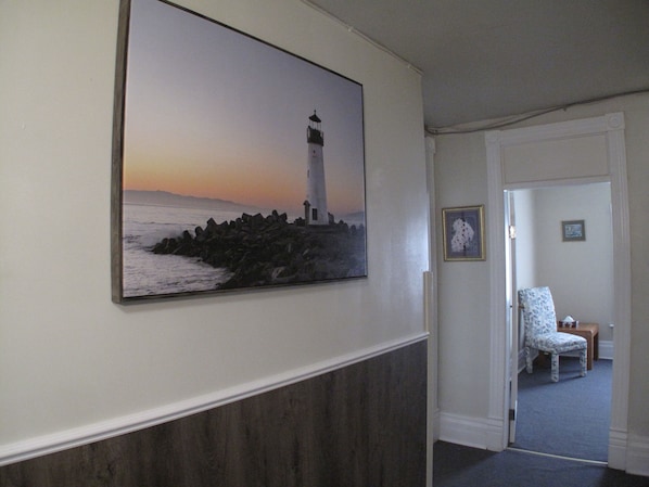 A hallway with a framed painting of a lighthouse, a patterned chair, and a door leading to another room.
