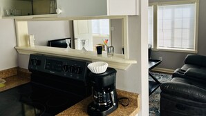 Eine kompakte Küche mit Herd, Kaffeemaschine und Mikrowelle. Im Wohnbereich befindet sich ein Fenster mit Jalousien und eine schwarze Ledersofa.