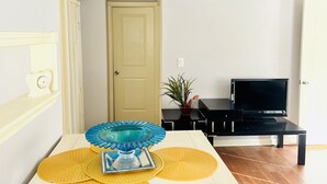 Ein Zimmer mit einer weißen Tür, einem schwarzen TV-Schrank, einem blau-gelben Tisch und einer Topfpflanze.