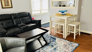 Ein Wohnzimmer mit einer schwarzen Ledersofa, einem kleinen schwarzen Couchtisch, einem grauen Sessel, einem weißen Esstisch mit zwei Stühlen und einem gemusterten Teppich.