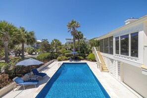 A rectangular swimming pool with a clear blue water, surrounded by lounge chairs and umbrellas, and a staircase leading to a building with large windows.