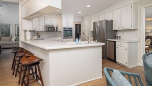 A modern kitchen with white cabinetry, stainless steel appliances, a central island with stools, and a view of the living area. - Spacious 4-Bedroom Beach House with Pool and Ocean Views (Isle Of Palms)
