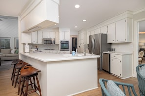 A modern kitchen with white cabinetry, stainless steel appliances, a central island with stools, and a view of the living area. - Spacious 4-Bedroom Beach House with Pool and Ocean Views (Isle Of Palms)