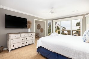 A bedroom with a large bed, a dresser, a flat-screen TV, and a view of the ocean through a sliding glass door.