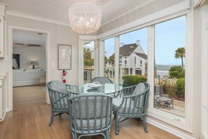 A dining area with a glass table and wicker chairs, overlooking a scenic view of a coastal house and beach.