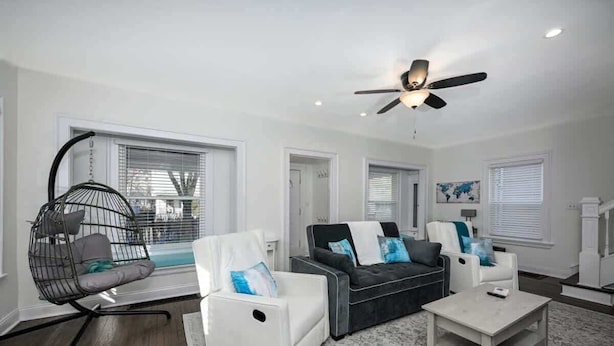 Una sala de estar moderna con un ventilador de techo, una silla colgante negra, un sillón blanco, un sofá gris con cojines azules y una mesa de centro.