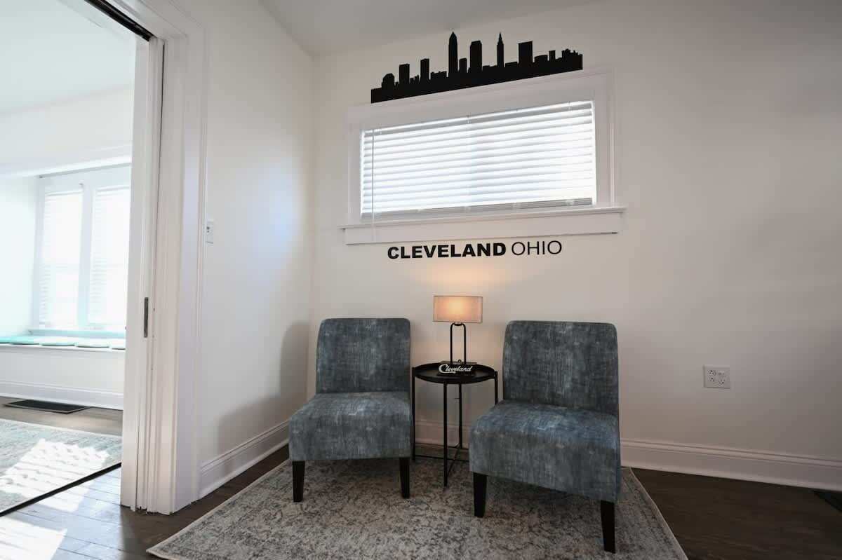 A hotel lobby with two grey armchairs, a small table with a lamp, a window with blinds, and a city skyline silhouette on the wall.