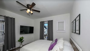 Un dormitorio con cama, ventilador de techo, televisión y ventana con cortinas.