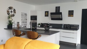 Una cocina moderna con una isla central, muebles blancos y encimera negra. Hay dos sillas de madera y un sofá amarillo en la zona de estar.
