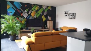 Una sala de estar moderna con un sofá amarillo, un televisor de pantalla plana y una zona de cocina con fregadero y encimera.