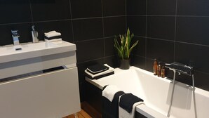 Un baño moderno con una bañera blanca, azulejos negros, un lavamanos con artículos de tocador y una planta en maceta.