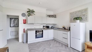 A modern kitchen with white cabinetry, a built-in oven, and a refrigerator. There is a fire extinguisher on the wall, a clock, and a map. - Beautiful 2-bed Apartment in Tunbridge Wells (Royal Tunbridge Wells)