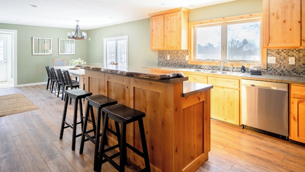 A kitchen with wooden cabinets, a central island with stools, a dishwasher, and a window providing natural light. - Private 5-Acre Estate | Fishing Pond + Fire Pit (West Olive)