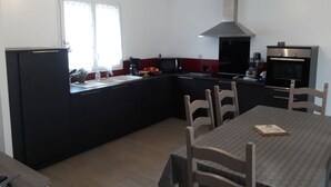 Een kleine keuken met een koelkast, fornuis, magnetron en wastafel. Er is een eettafel met stoelen en een raam met gordijnen.