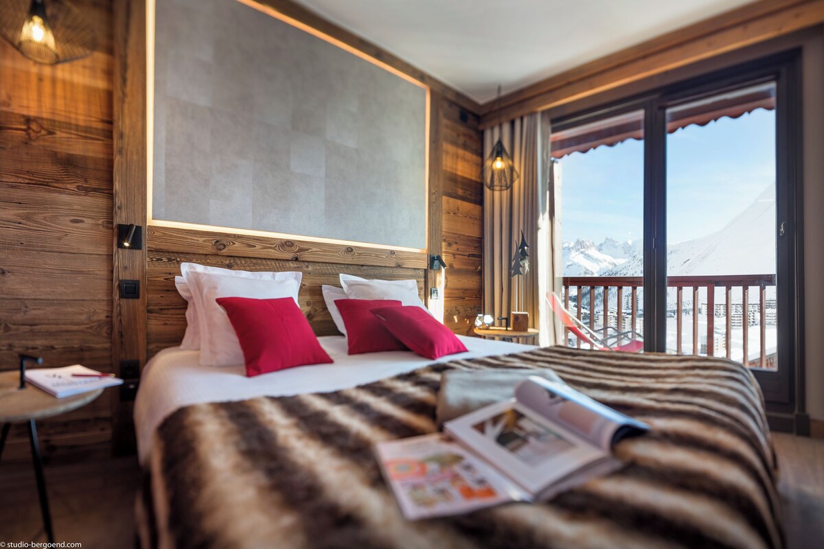 Habitación de hotel con una cama grande, paredes de madera, un balcón con vistas a las montañas y una ventana con cortinas.