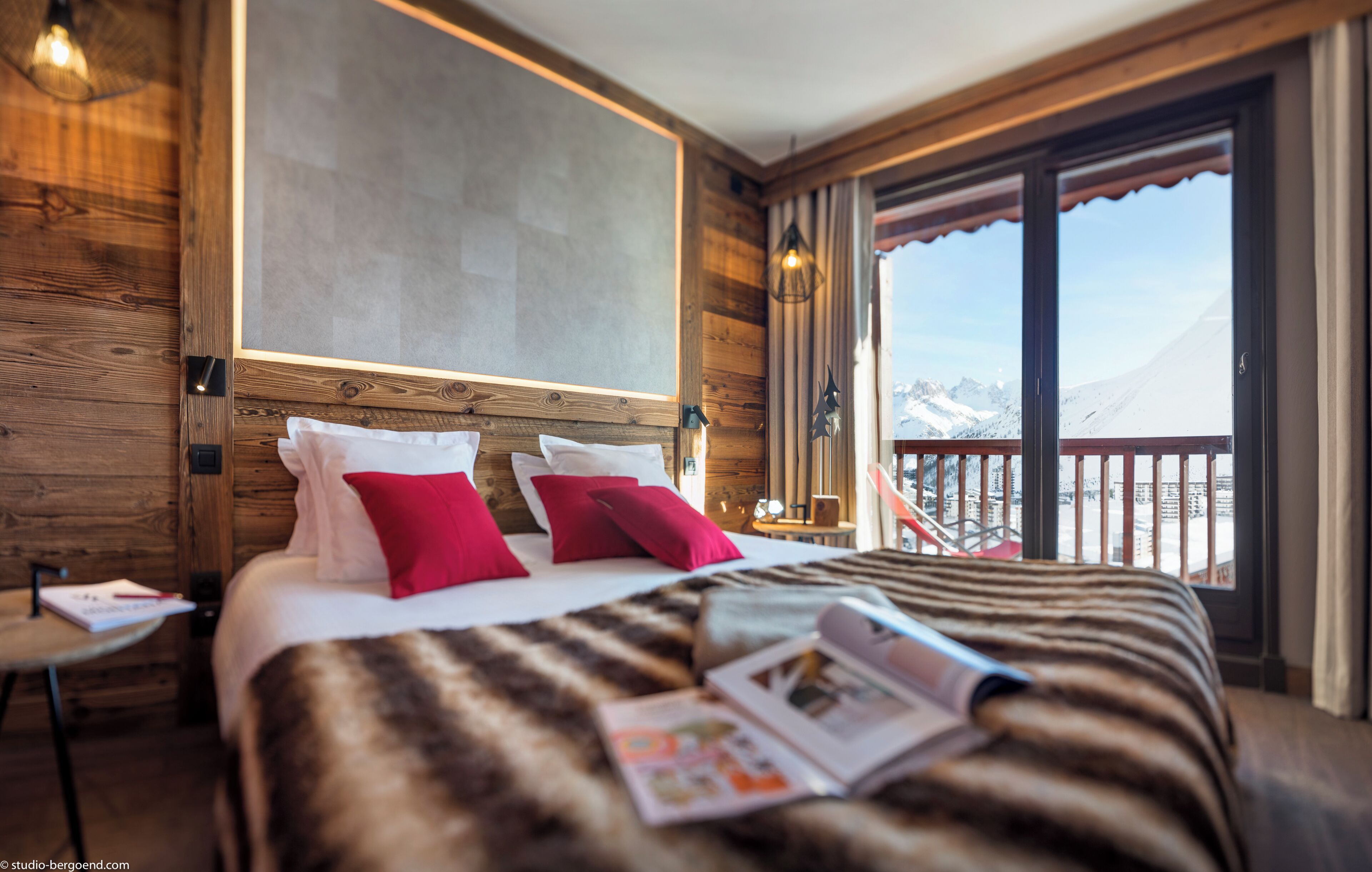 A hotel room with a large bed, wooden walls, a view of mountains, and a balcony with a railing.
