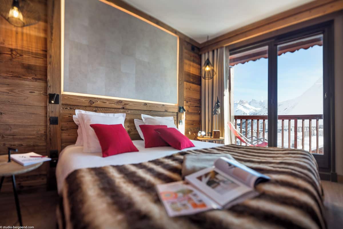 Ein Hotelzimmer mit einem großen Bett, Holzwänden, einem Balkon mit Bergblick und einem Fenster mit Vorhängen.