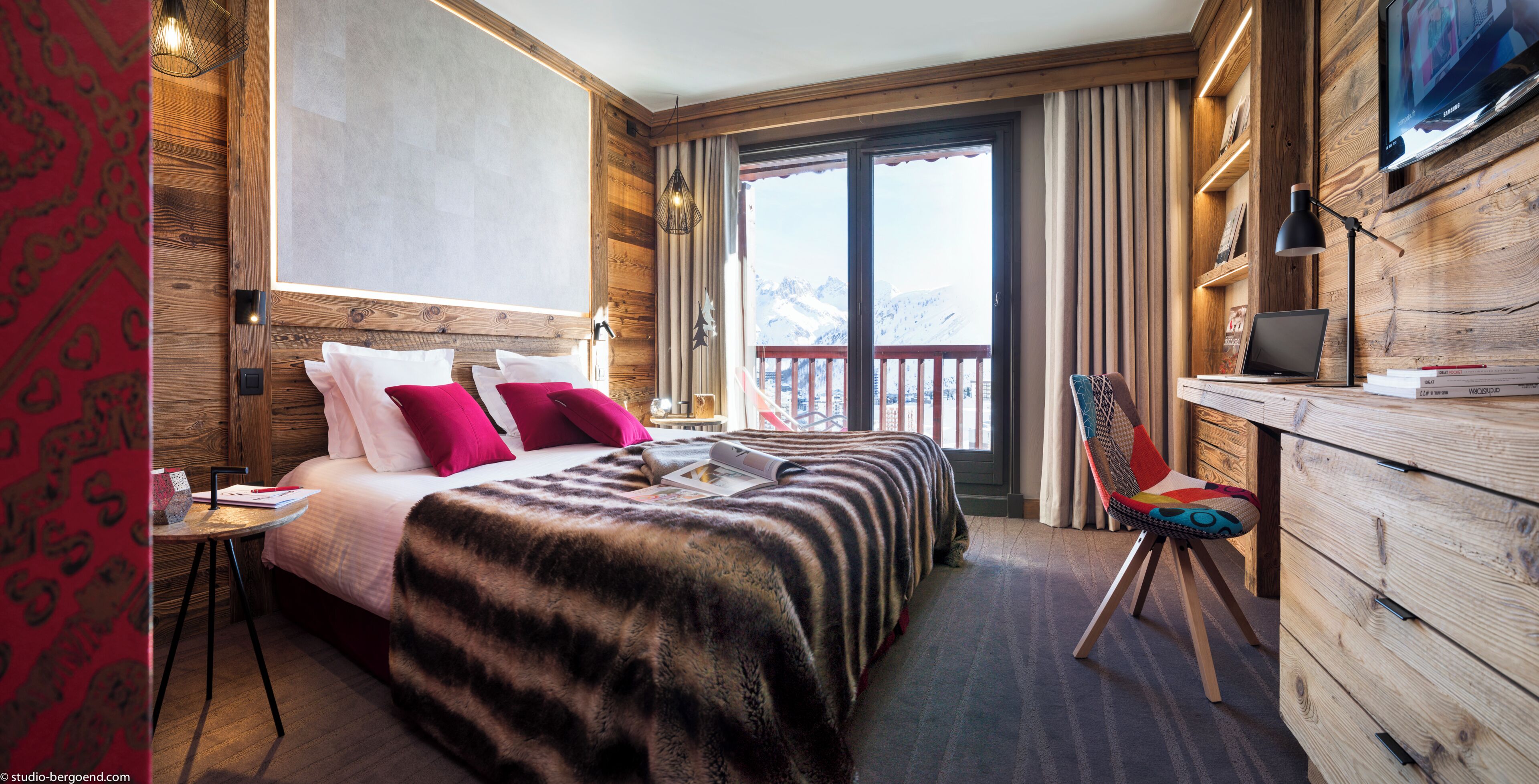 A hotel room with a bed, a desk with a laptop, a chair, and a view of mountains.