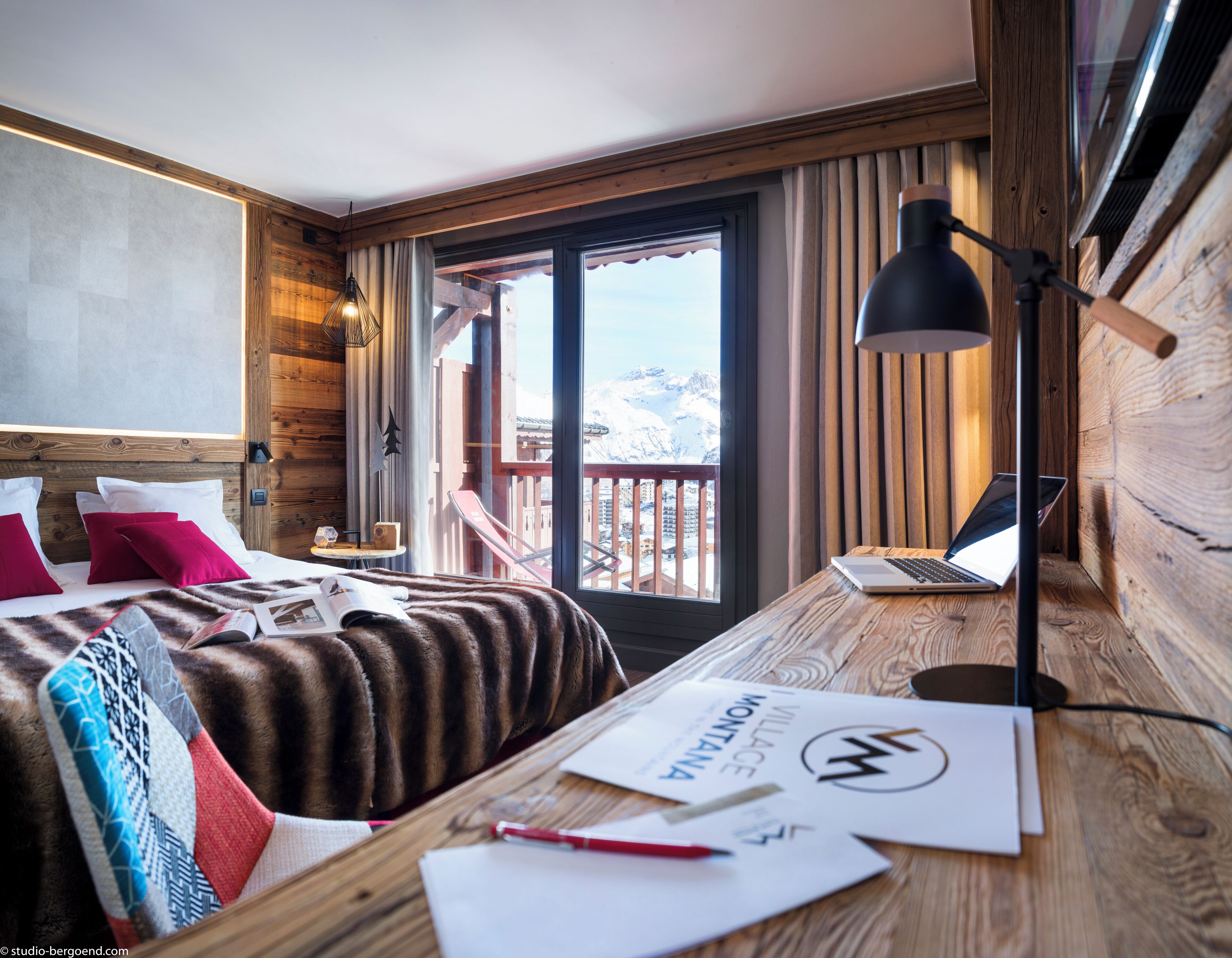 A wooden room with a bed, a desk with a laptop, and a view of mountains.