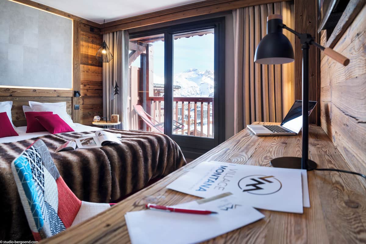Ein Zimmer mit Holzboden, einem Bett, einem Schreibtisch mit einem Laptop und Blick auf Berge durch ein Fenster.