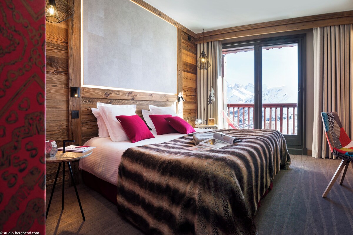Habitación de hotel con cama, escritorio con ordenador portátil, silla y vista a las montañas a través de la ventana.