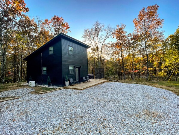 A modern two-story building with a dark exterior, surrounded by a gravel driveway and a wooden deck with outdoor seating.
