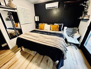 A bedroom with a bed, bedside table, and a mirror. There is a wall-mounted air conditioner and a window with a view of the outdoors.