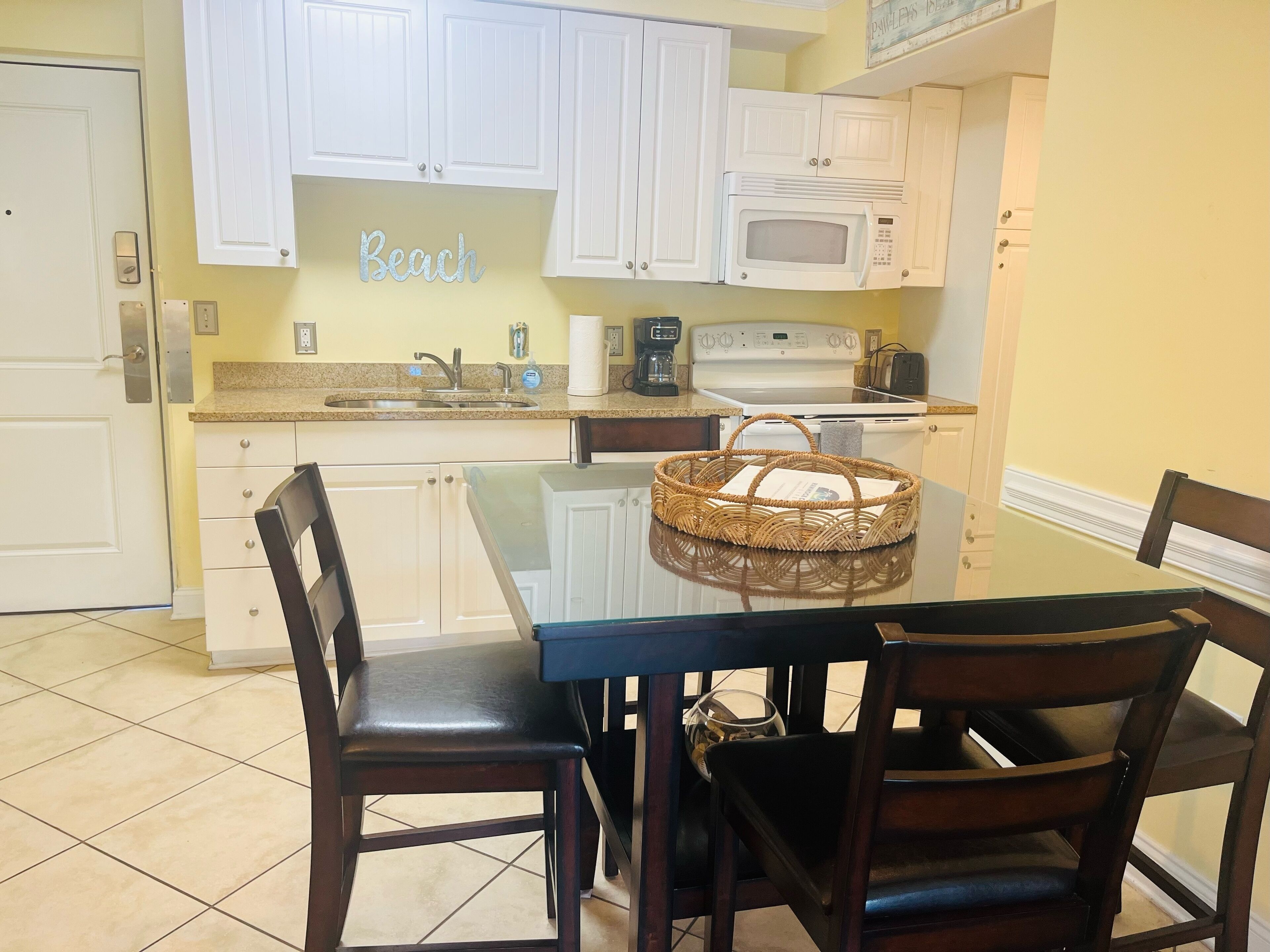 A compact kitchen with white cabinets, a microwave, a coffee maker, and a glass-top dining table with chairs.
