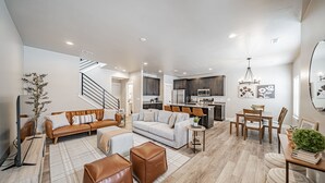 A spacious living area with a sofa, ottoman, and a dining table with chairs. There is a staircase leading to another level, and a kitchen area with a microwave and cabinets.