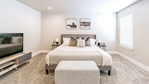 A bedroom with a large bed, a TV on a stand, a bedside table, and a window with blinds.