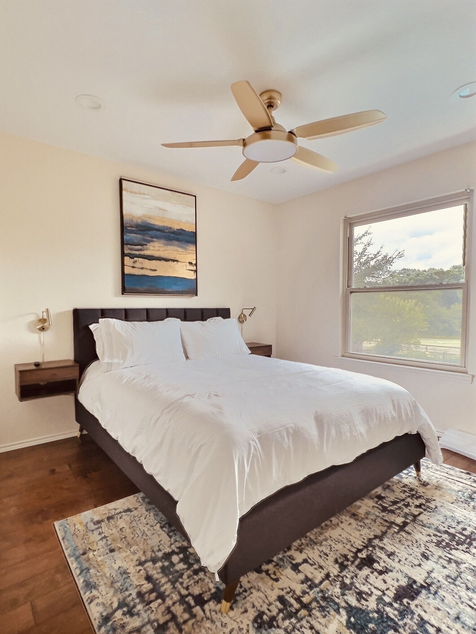 A bedroom with a bed, a ceiling fan, a painting, a nightstand, and a window.