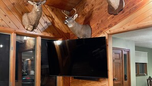 A room with wooden walls and ceiling, a mounted deer head on the wall, a mounted bear head, and a large flat-screen TV. - Cozy Lodge (Sherman)